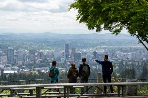 Four people stand on a viewpoint overlooking a city skyline, with green trees and mountains in the background.