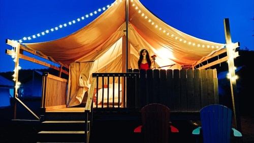 A woman stands on a deck of a glamping tent, illuminated by string lights, with a cozy atmosphere at dusk.