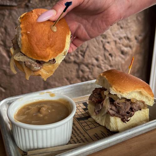 A hand holds a beef sandwich with onions, next to another sandwich and a bowl of gravy on a metal tray.