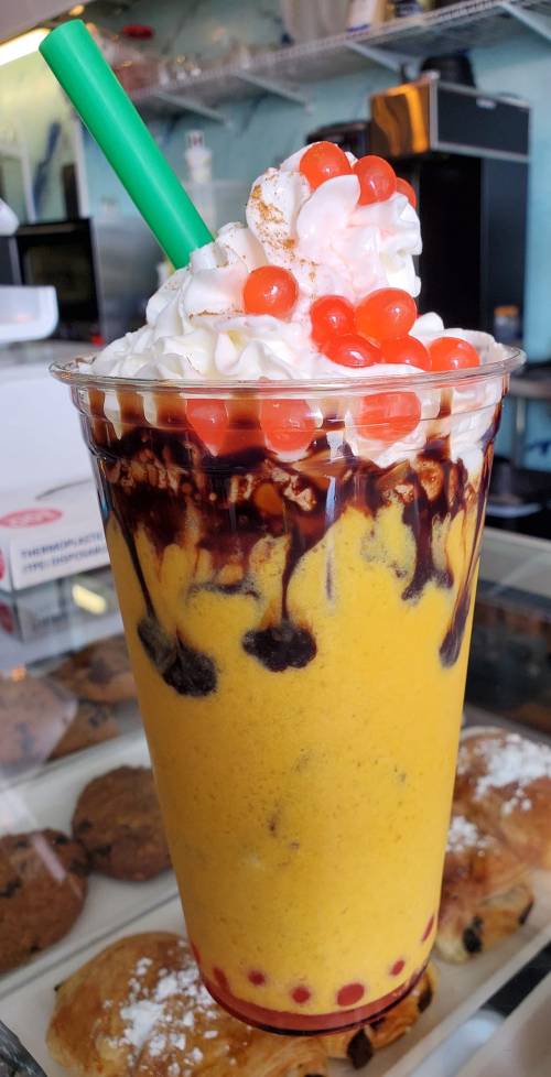 A colorful smoothie topped with whipped cream, red jelly beads, and chocolate drizzle in a clear cup with a green straw.