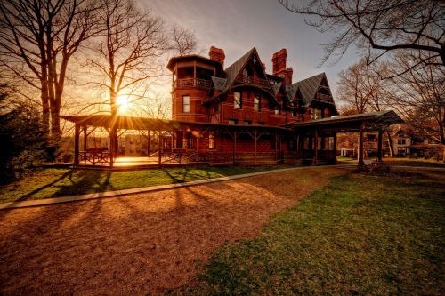 A historic red mansion at sunset, surrounded by trees and a grassy area, with a covered porch in the foreground.