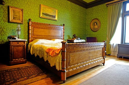 A vintage bedroom with a wooden bed, green patterned walls, a nightstand, and framed artwork.