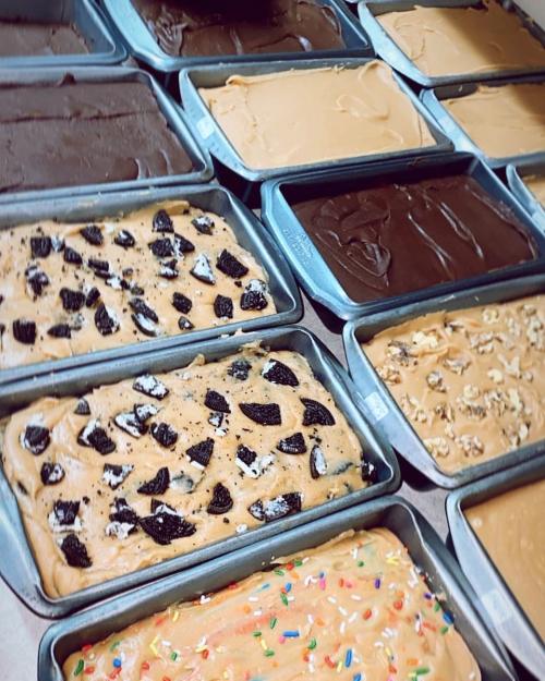 A variety of fudge in metal trays, featuring flavors like chocolate, peanut butter, and topped with cookies and sprinkles.