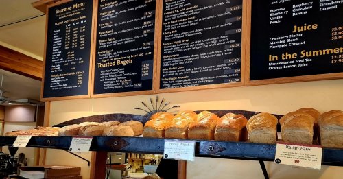 A café menu board with espresso options and a display of various bread loaves on a shelf below.