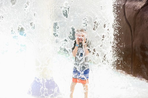 A child stands playfully in a splash of water, surrounded by a cascade of droplets at a water park.