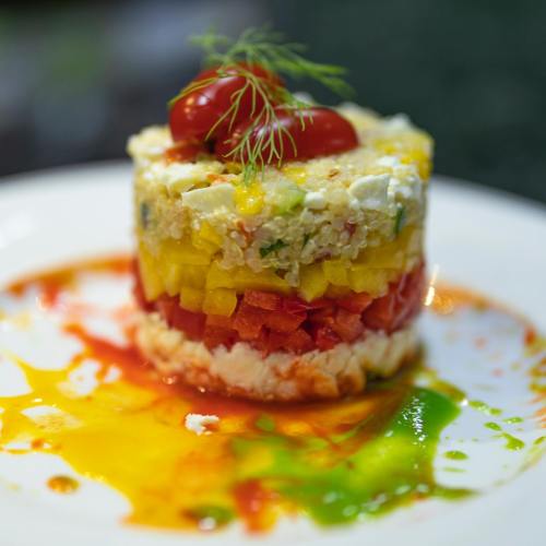 A colorful layered salad with diced vegetables, cheese, and cherry tomatoes, drizzled with vibrant sauces on a plate.