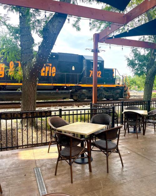 A train passes by a café patio with empty tables and chairs under shaded awnings.
