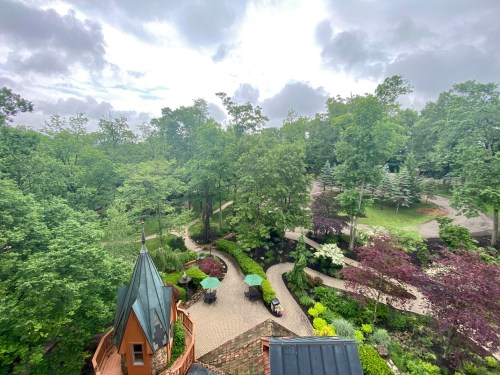 Aerial view of a lush green garden with winding paths and a unique building in the foreground under a cloudy sky.