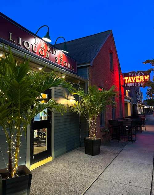 Exterior of a liquor store and tavern at dusk, featuring palm plants and illuminated signs.