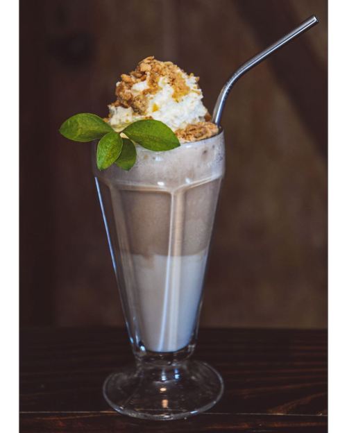 A tall glass of milkshake topped with whipped cream, crushed cookies, and a green leaf, served with a metal straw.