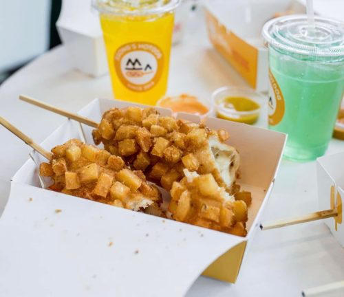 Two crispy potato-covered hot dogs on skewers in a takeout box, with drinks and sauces in the background.