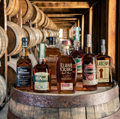 A collection of whiskey bottles displayed on a barrel in a rustic distillery setting, surrounded by wooden barrels.