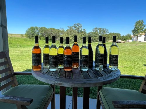 A table with various bottles of wine arranged in two rows, set against a clear blue sky and green landscape.