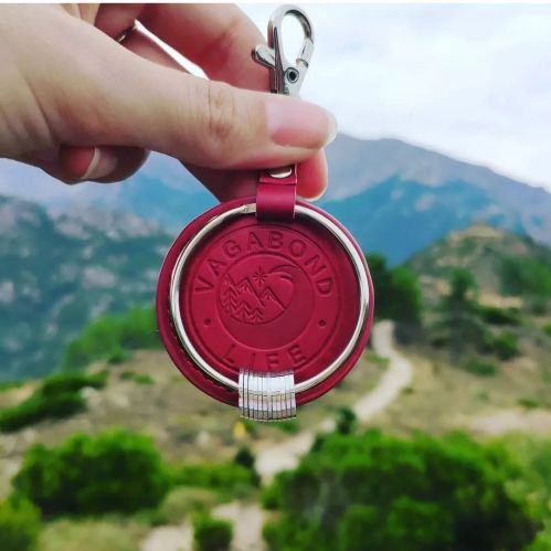 A hand holds a red keychain with "VAGABOND LIFE" embossed, set against a scenic mountain backdrop.
