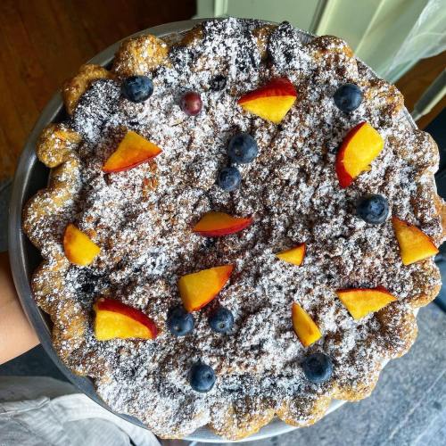A round dessert topped with powdered sugar, blueberries, and peach slices on a silver platter.