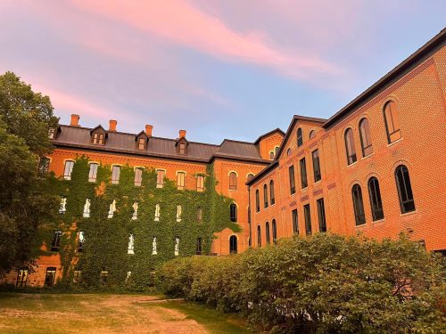 Historic brick buildings covered in green ivy, set against a colorful sunset sky.