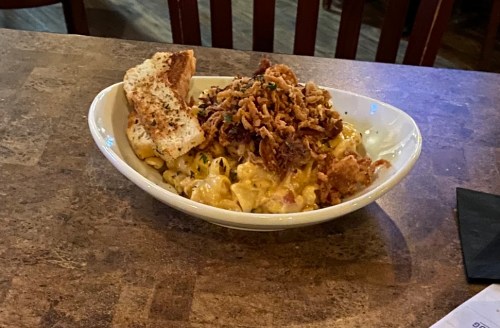 A bowl of creamy macaroni and cheese topped with crispy onions and a slice of toasted bread on a wooden table.