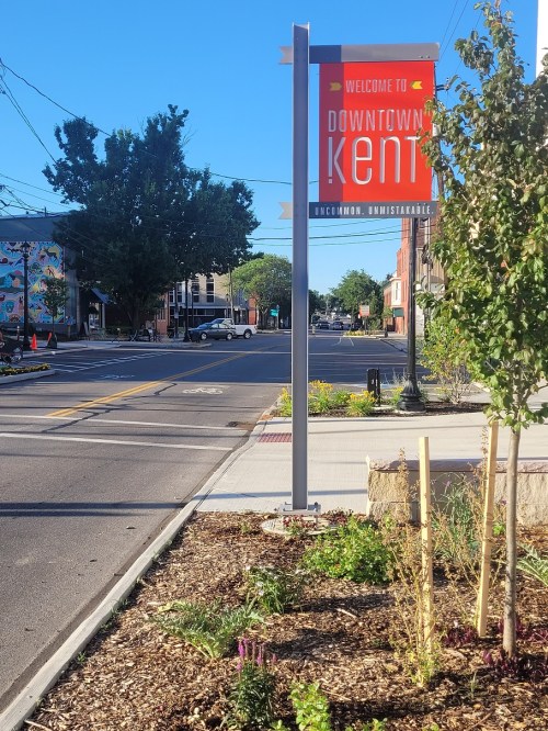 A welcome sign for Downtown Kent stands beside a landscaped area, with a view of the street and buildings in the background.