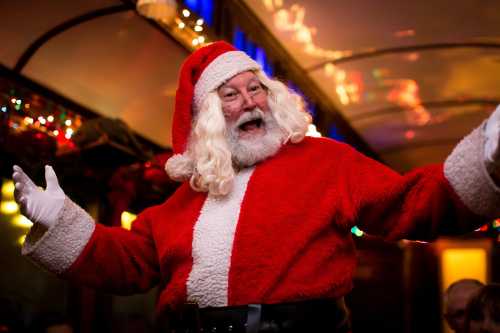 A joyful Santa Claus in a red suit and hat, smiling and gesturing cheerfully in a festive setting.