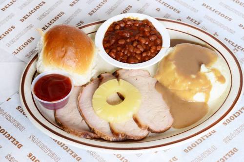 A plate with sliced pork, pineapple, mashed potatoes with gravy, baked beans, and a roll, served with ketchup.