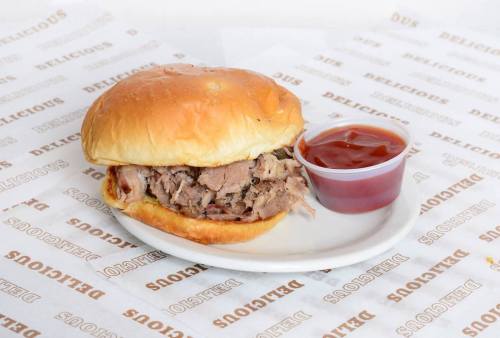 A pulled pork sandwich on a soft bun, served with a side of barbecue sauce in a small cup.