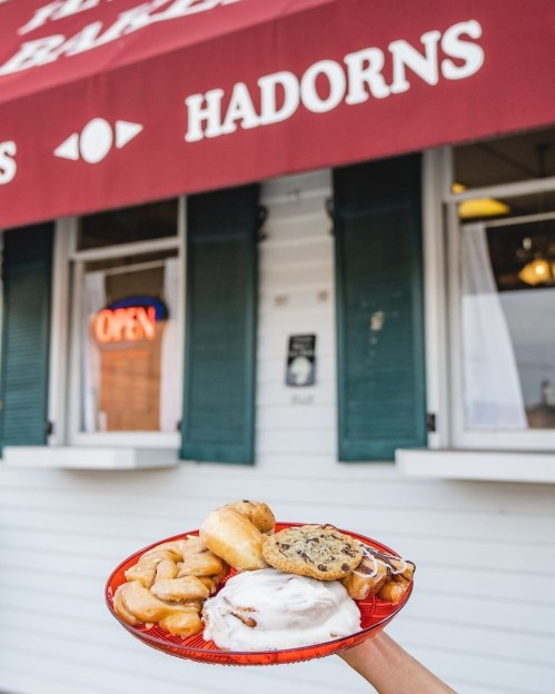 A hand holds a red plate with pastries, a cinnamon roll, and a cookie in front of a bakery with an "Open" sign.