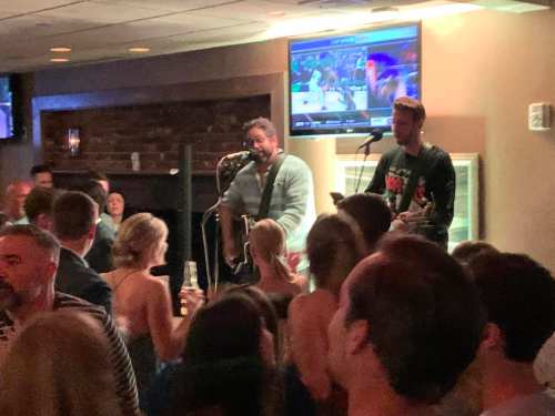 A lively bar scene with two musicians performing on stage while a crowd enjoys the music and drinks.