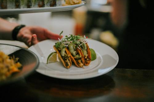 A plate of three tacos topped with greens and served with lime wedges, held by a person in a restaurant setting.