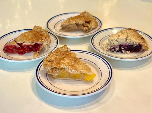 Four slices of pie on white plates, featuring different fillings: cherry, blueberry, peach, and a mixed fruit pie.