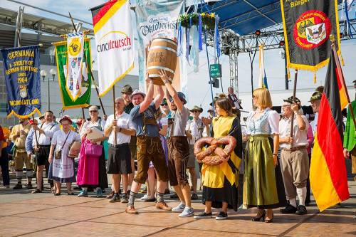 A festive group in traditional attire celebrates at an outdoor event, holding flags and a large beer barrel.