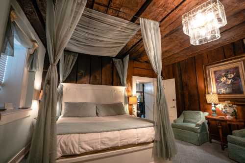 Cozy bedroom with a four-poster bed, soft curtains, a chandelier, and a vintage armchair in a rustic setting.