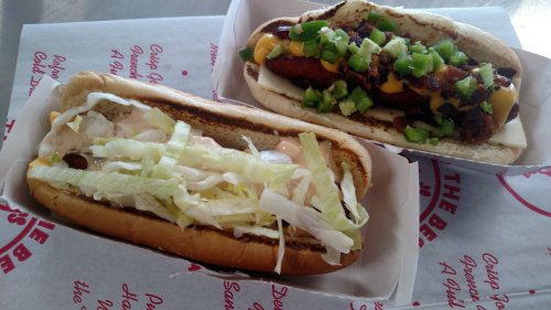 Two hot dogs in buns: one topped with lettuce and sauce, the other with cheese, bacon, and chopped green onions.