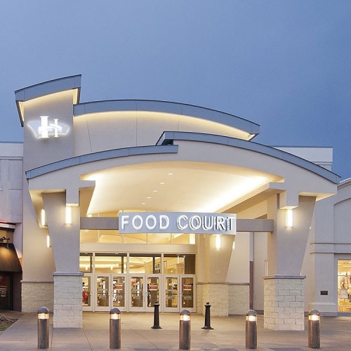 A modern food court entrance with a curved roof and bright signage, surrounded by outdoor seating and light posts.
