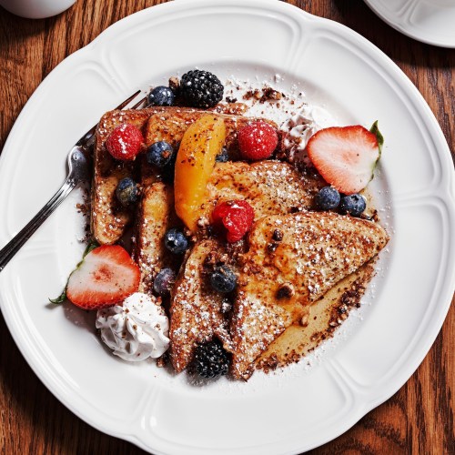 A plate of French toast topped with fresh berries, peach slices, and whipped cream, dusted with powdered sugar.