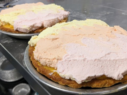 Two pies with a thick layer of pastel-colored whipped cream on top, resting on metal trays.