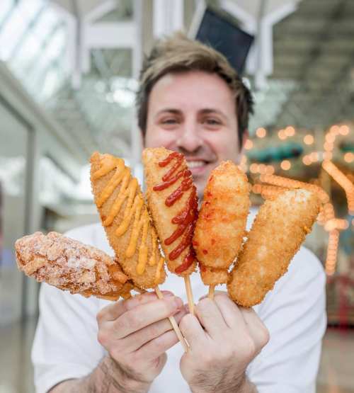 A person holds five different corn dogs on sticks, showcasing various toppings and coatings in a festive setting.