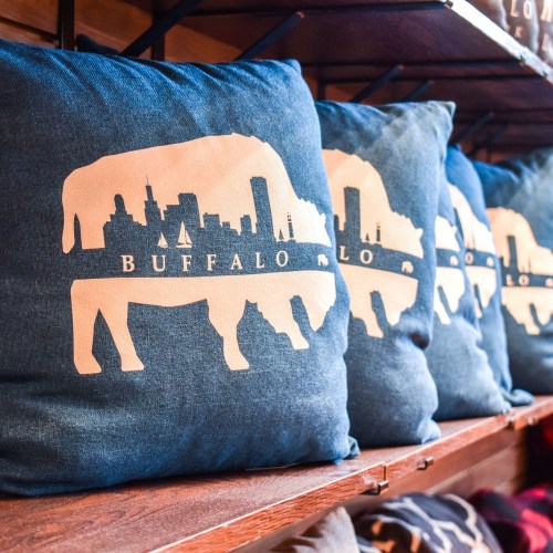 Three decorative pillows featuring a silhouette of a buffalo with a city skyline design, displayed on a wooden shelf.