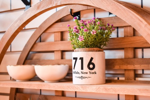 A wooden shelf displays a white pot with flowers labeled "716 Buffalo, New York" and two small bowls.
