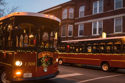 Two festive trolleys decorated with lights, one labeled "North Pole," parked on a city street at dusk.