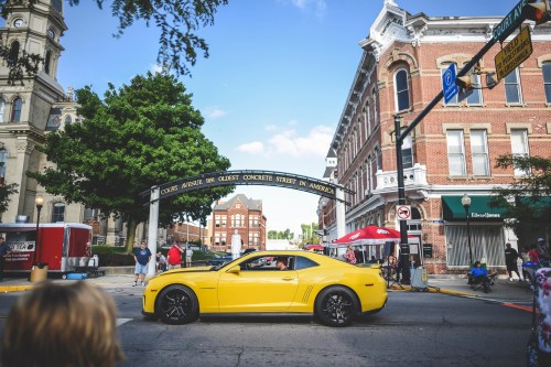 A yellow sports car drives through a lively street scene with historic buildings and people enjoying the day.