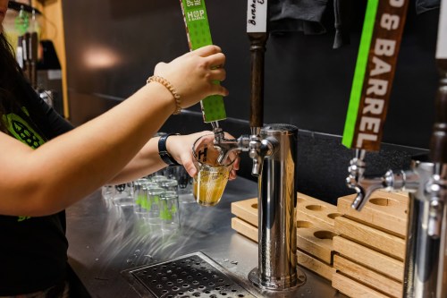 A person pours a drink from a tap into a clear glass at a bar, with wooden coasters in the background.