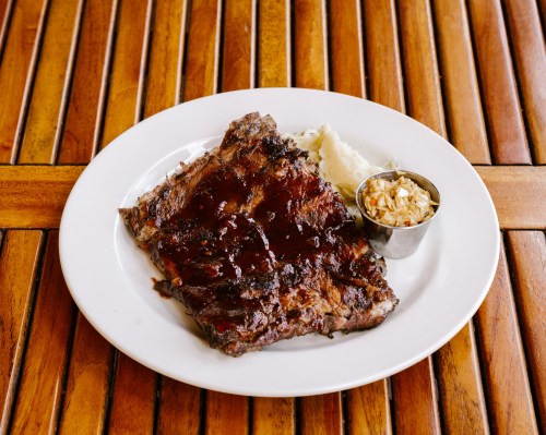 A plate of barbecue ribs with a side of mashed potatoes and coleslaw, served on a wooden table.