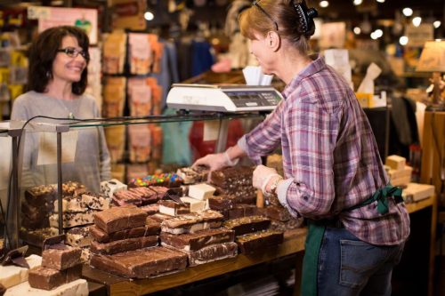 A woman serves fudge and sweets at a counter while a customer smiles in a cozy candy shop.