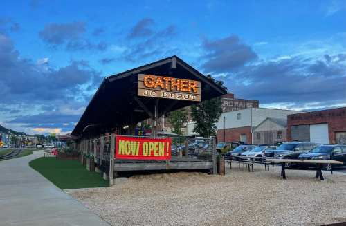 A rustic outdoor gathering space with a "NOW OPEN!" banner, surrounded by cars and under a blue sky.