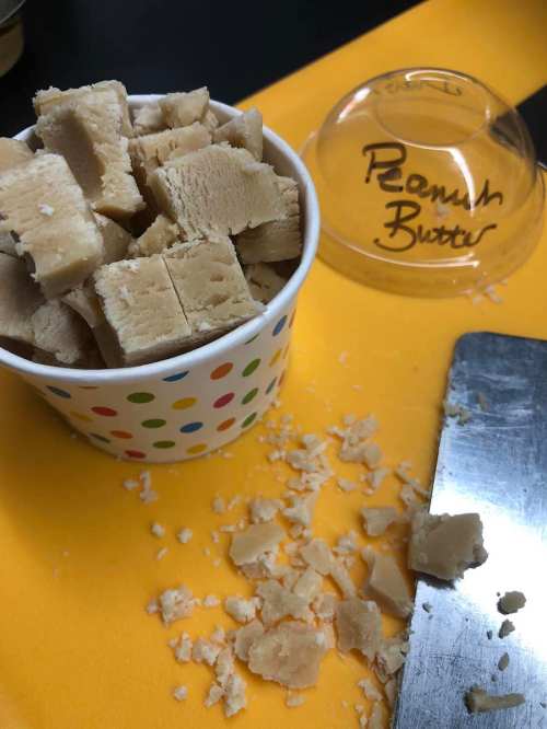 A cup of cubed peanut butter fudge with some pieces scattered on a yellow surface.