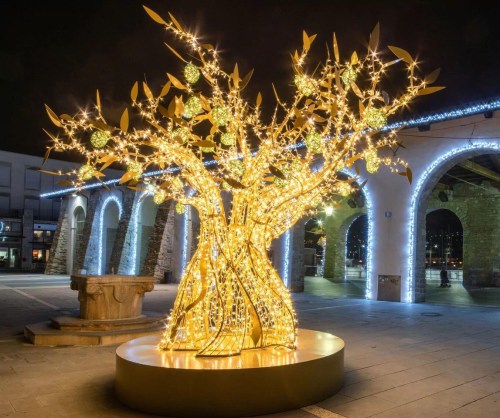 A glowing, artistic tree made of lights and ornaments, set in a nighttime urban plaza.