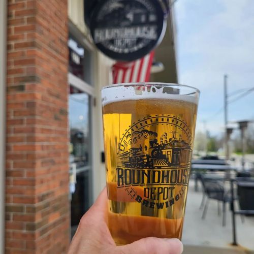 A hand holds a pint of beer in front of a brewery with a sign and an American flag in the background.