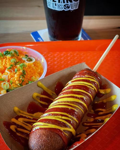 A corn dog drizzled with ketchup and mustard, served with a side of orange rice and a glass of dark beer.