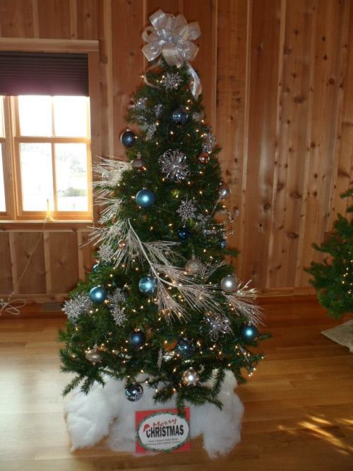 A decorated Christmas tree with blue and silver ornaments, lights, and a bow, set against a wooden wall.
