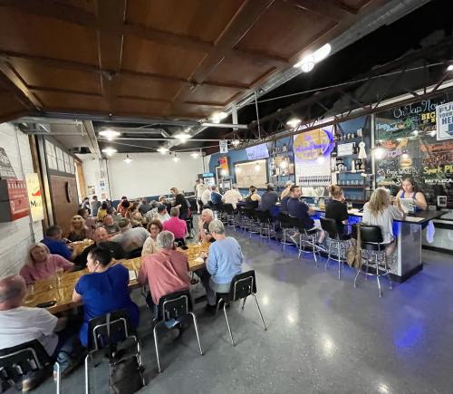A lively restaurant scene with diners at long tables and a bar, featuring a vibrant mural and modern decor.